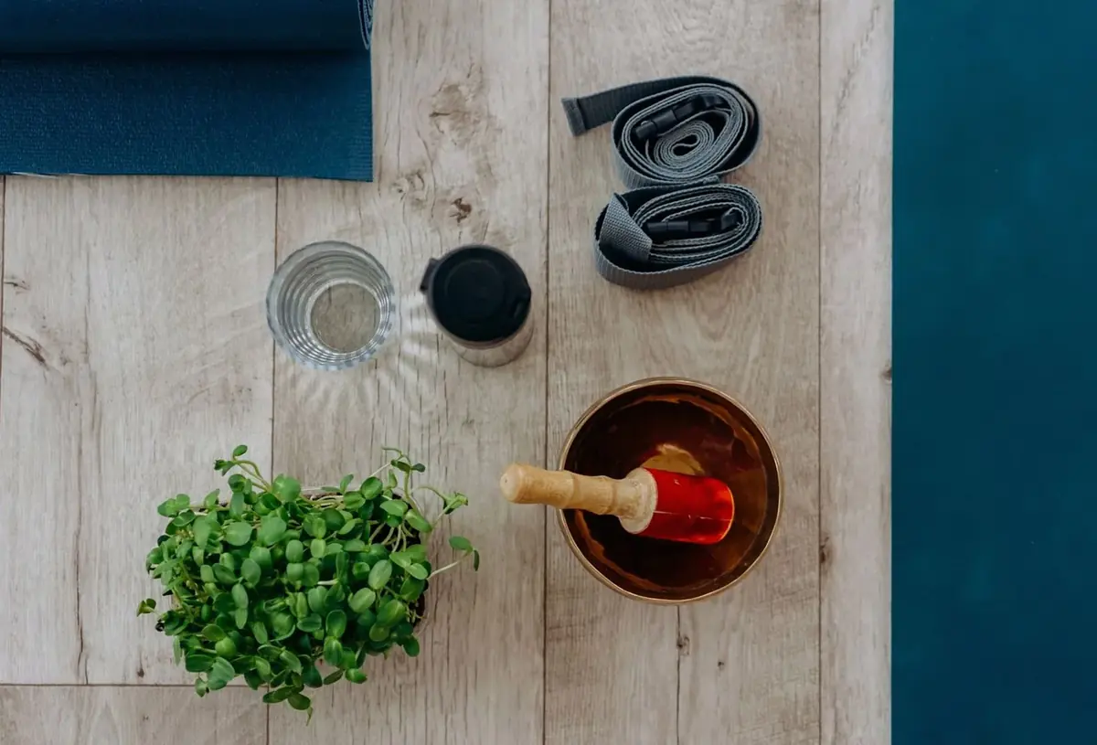 Yoga mat, strap, glass of water, singing bowl and a small potted herb on a wooden floor